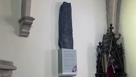 Oruths cross is mounted to a church wall. It's resting on a podium which is titles "Orsuths Cross"