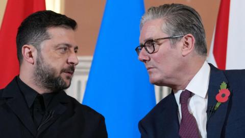 Starmer leans in to talk to Zelensky - they're standing in front of a red, blue and white background
