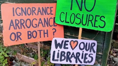 Three colourful protest signs lean against a green fence. One reads "ignorance, arrogance or both"; one reads "no closures"; and one reads "we love our libraries", with the word "love" being represented by a pink love-heart. 