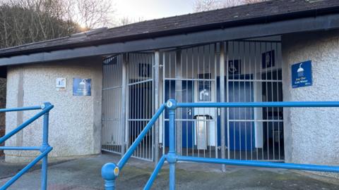 A public toilet block with its entrance blocked by silver steel gates and bars. There are blue metal railings in front of the building, which has off-white textured walls and blue signs for showers and men, women and baby changing facilities. 