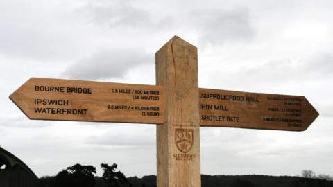 A wooden fingerpost with Bourne Bridge, Ipswich Waterfront, Suffolk Food Hall, Pin Mill and Shotley Gate written on it. In the middle there is also the St Joseph's College emblem.