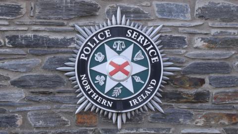 The official badge of the Police Service of Northern Ireland is displayed on a grey brisk wall.
