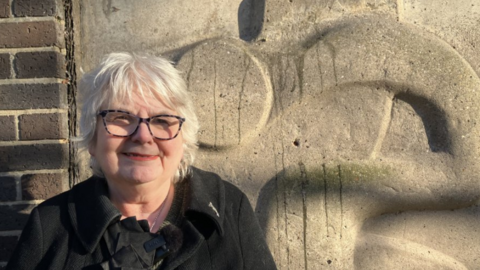 Angela Greenfield - a woman with short, grey hair, smiling. She is wearing dark glasses and a black coat. She is standing in front of a stone-coloured sculpture which is on the side of a car park