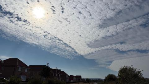 A picture of the blue sky showing a dense clumped-together cloud pattern which stretches across the image. At the bottom there are the dark silhouettes of houses and trees, and the sun is shining through the white fluffy clouds.