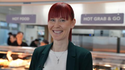 Lorraine Walker is standing in a school canteen. She is wearing a green blazer and white top. She has red hair.