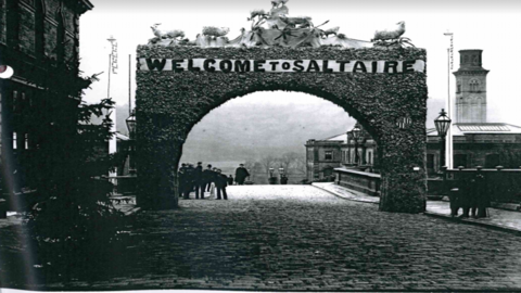 An old black and white photo shows a huge decorative arch made of flora and foliage, with the word 'Saltaire' across the top in large letters. It sits in a cobbled street, and mill style buildings and a tower in the background in indicate this is an industrial area. Figures in the background wear bowler hats and clothing which looks late 19th Century in style.