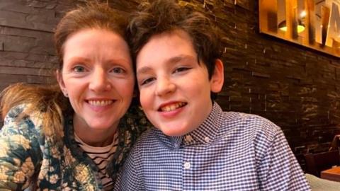 Kelly and her son are sat together in front of a restaurant wall. Kelly on the left with light brown hair and smilling, in a flowery jacket. Gael has a blue checked shirt on, is smiling and has dark brown hair.