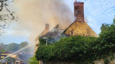 Stone building with fire engines parked in front and water from a firefighter's hose spraying onto the building.