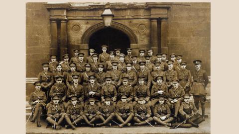 Photo of 48 men outside an archway in military outfits. The men are posing for a class photo. There is a lantern above them. The photo is sepia-toned. 