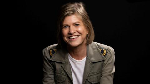 Clare has short dark hair in a bob with a side parting. She smiles for a portrait wearing a white t shirt and a military-style jacket over it.