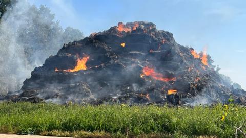 A view of the burning stack of hat. It has been burnt black and fire can be seen in several parts of the pile. Smoke billows out.