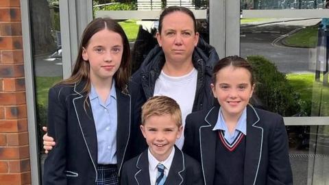 Rebecca with her three children, who are all wearing school uniforms, standing outside the entrance to Wellington School