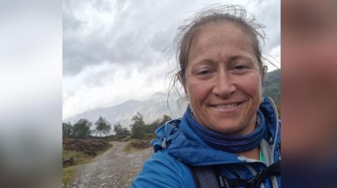 A selfie of Rachel Morris wearing a blue coat and blue snood with a misty mountain scene in the background. She is looking at the camera