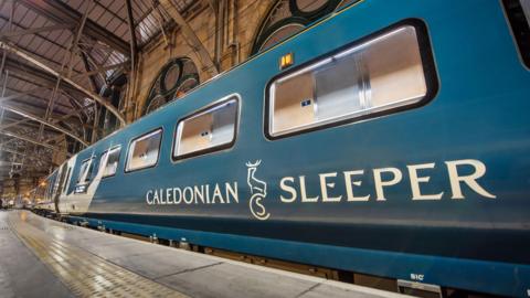 A dark blue train carriage is photographed at a train station. The carriage has white patterns across it and "Caledonian Sleeper" written on the side in white writing. 