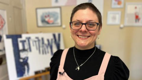 Kate Rolfe, she has short dark hair, is wearing a black top with pink apron, behind her are artworks. 