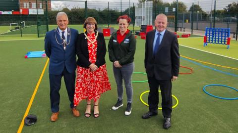 A number of people standing on one of the new sports pitches, There is a variety of equipment, including hoops and plastic barriers. The pitch has flood lights and is surrounded by a high mesh fence. 