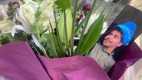 A close up of a bouquet of purple and white flowers wrapped in purple paper with a picture of Mohammed Algasim.