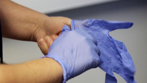 A picture of someone putting on blue gloves. They are stood in a room in front of a white wall.