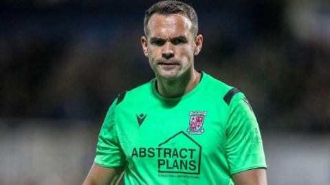 Craig Ross wears a green goalkeeping jersey with a Woking badge