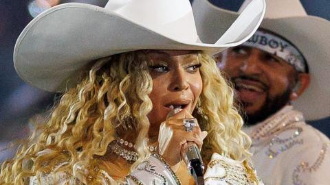 Beyonce in a white cowboy hat and jewelled white suit singing into a microphone flanked by dancers in similar outfits, performing at half-time furing an NFL game on 25 December 2024