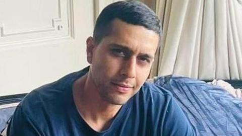 Llun o Kamran Aman. Mae'n gwisgo crys T glas ac yn edrych i fewn i'r camera.