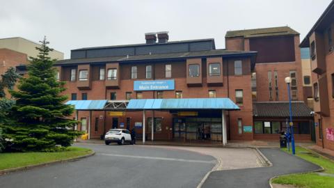 The exterior of Scarborough Hospital, a brick building of three stories with a blue canopy at the front, a driveway with a white car parked on it and a fir tree to the side.