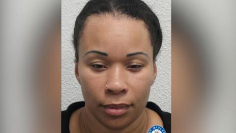 A woman with pulled-back black curly hair looking at the camera in a police mugshot. She is wearing a black top and small gold nose ring.