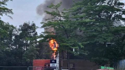 Flames from a building surrounded by metal fencing, with lots of smoke wafting above the treeline