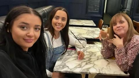 Louise, Hannah and Carol Hunt sit around a table, holding drinks and smile into the camera