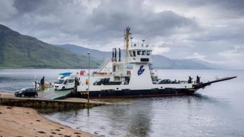 A car and a campervan disembark from the ferry MV Corran. The ferry has Highland Council's logo on its side. The logo is in the shape of the Highland Council area.