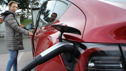 A woman stands by a Tesla model Y as it charges. 