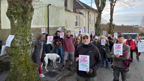 People in coats holding up signs that say "Stop the Chop". There are trees next to them. To the left is a yellow house. Behind them are cars. The sky is blue with white clouds. 