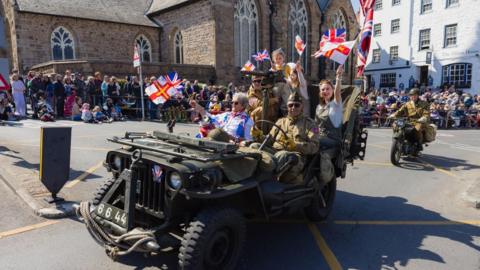 People sat in a green vehicle. They are waving Guernsey flags and there is a crowd of people around them. There is a motorbike following the vehicle.