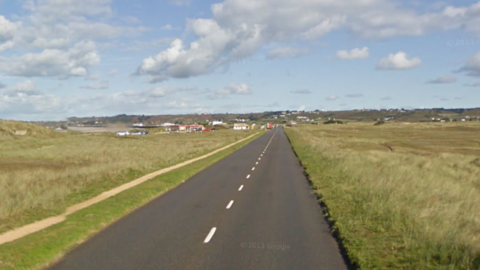 A  view along La Route de la Pulente. it is a long straight road with grass either side of it. There are cars on the road in the distance. 