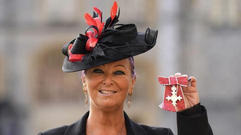 Charlotte Charles, wearing a black red hat, has purple tied-back hair and dangly earrings. She is holding an MBE medal in her right hand. She is wearing a black jacket and a necklace.