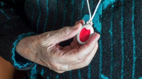 A elderly woman's hand holding a red and white plastic personal alarm device, worn round her neck on a white cord.