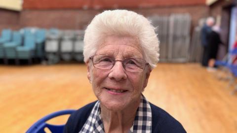 Jean Gooden is wearing glasses and is sitting down in a sports hall of Kings Heath Community Centre