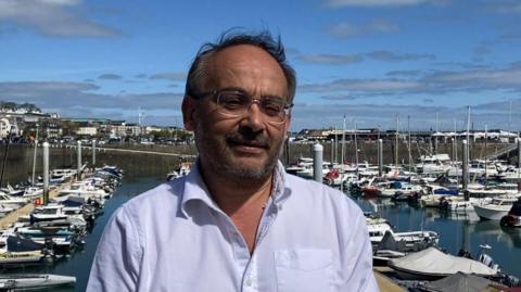 Deputy Chris Blin is pictured in front of a harbour scene. He is wearing a light blue shirt and has dark wispy hair with glasses and a short beard. 