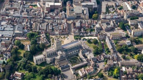 An aerial shot of Gloucester city centre on a sunny day, including Gloucester Cathedral in the foreground, green spaces, Georgian buildings, and Shire Hall