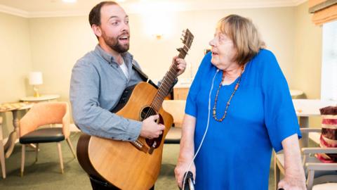 A man playing a guitar while looking at a woman, both have their mouths open in song.