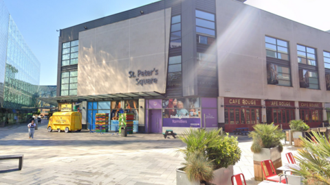 An open, concrete floor space with restaurant table and chairs to the bottom right. A grey building with black-framed buildings is in the background.