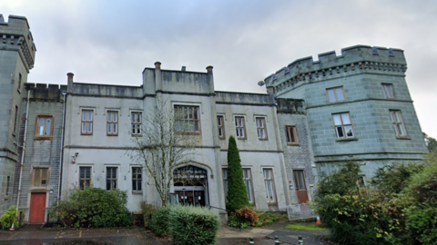 A large building, with various greenery in front of it. The building is a large, regal structure, possibly a redeveloped castle, including what looks like a tower as part of it.