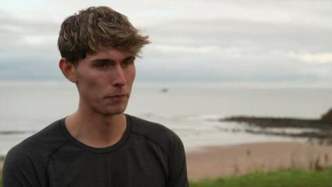 Leodhais Macpherson, in a black long-sleeved running t-shirt, stands with his back to the sea. He has dark blond hair.