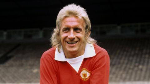 Denis Law smiles wearing a red Manchester United strip.