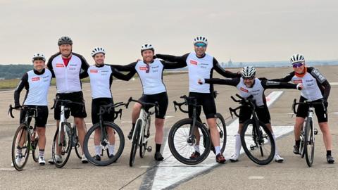 A team of seven male cyclists in black and white clothing standing astride their bikes on a runway of an airport. 