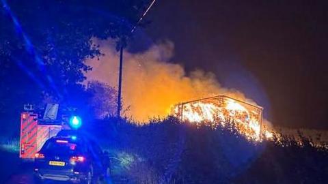 Bright orange flames rise from the frame of a large building, with smoke drifting into the night sky. To the left of the burning barn is a fire engine, lights flashing, and another vehicle