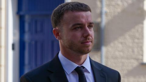 A man wearing a black suit and tie walking towards a building. Blue door and cream building behind.