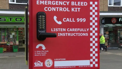 A red and white emergency bleed control kit fixed to a lamppost in a street.