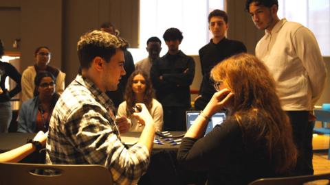 Two people engaged in conversation being watched by a group of young people