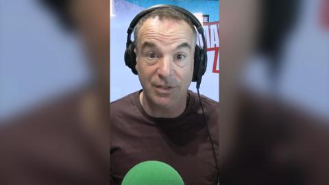 Martin Lewis wears headphones and a brown t-shirt. He sits in-front of a green microphone and looks directly at the camera.